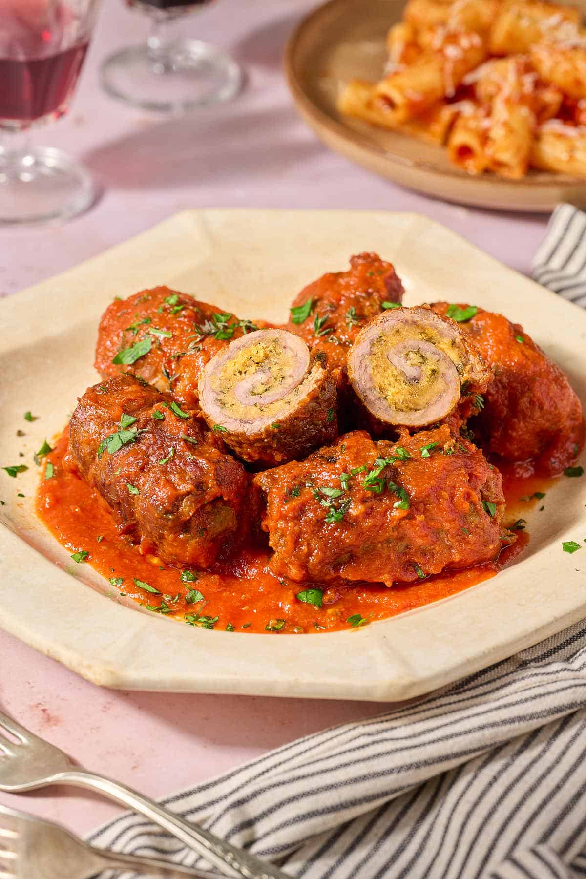 Pieces of braciole on a plate. Next to this is a cloth napkin, 2 forks, a glass of red wine and a plate of pasta with marinara sauce.
