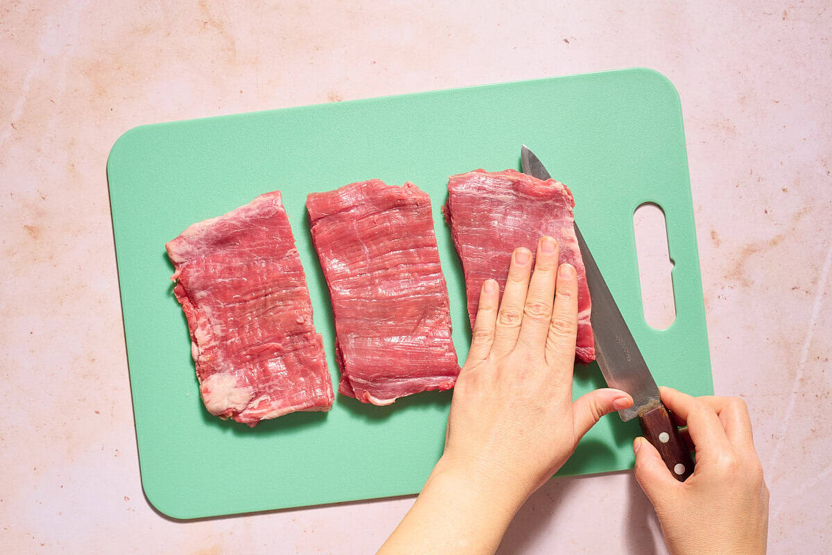 The flank steak for the braciole being cut into cutlets on a cutting board with a knife.