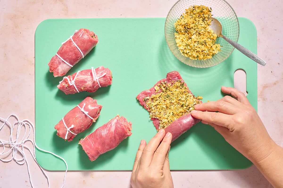 A single beef braciole being rolled up on a cutting board next to 4 other rolled braciole and the bowl of filling with a spoon on a cutting board.