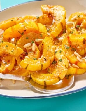Roasted delicata squash topped with pine nuts on a platter with a serving spoon.