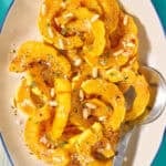 Roasted delicata squash topped with pine nuts on a platter with a serving spoon.