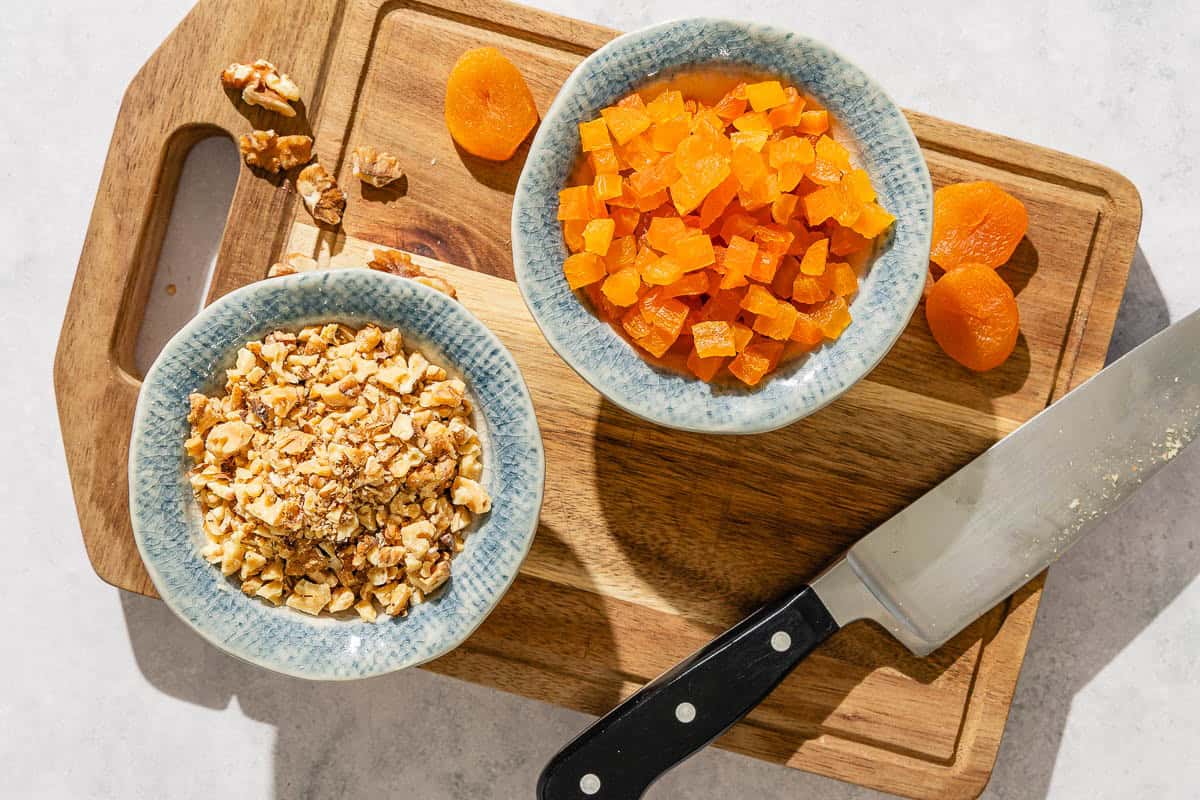 A bowl of chopped dried apricots and a bowl of chopped walnuts on a cutting board with a knife.