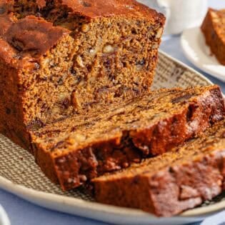 A partially sliced loaf of date nut bread behind 2 slices of the bread on a platter. Next to this is a slice of the bread on a plate.