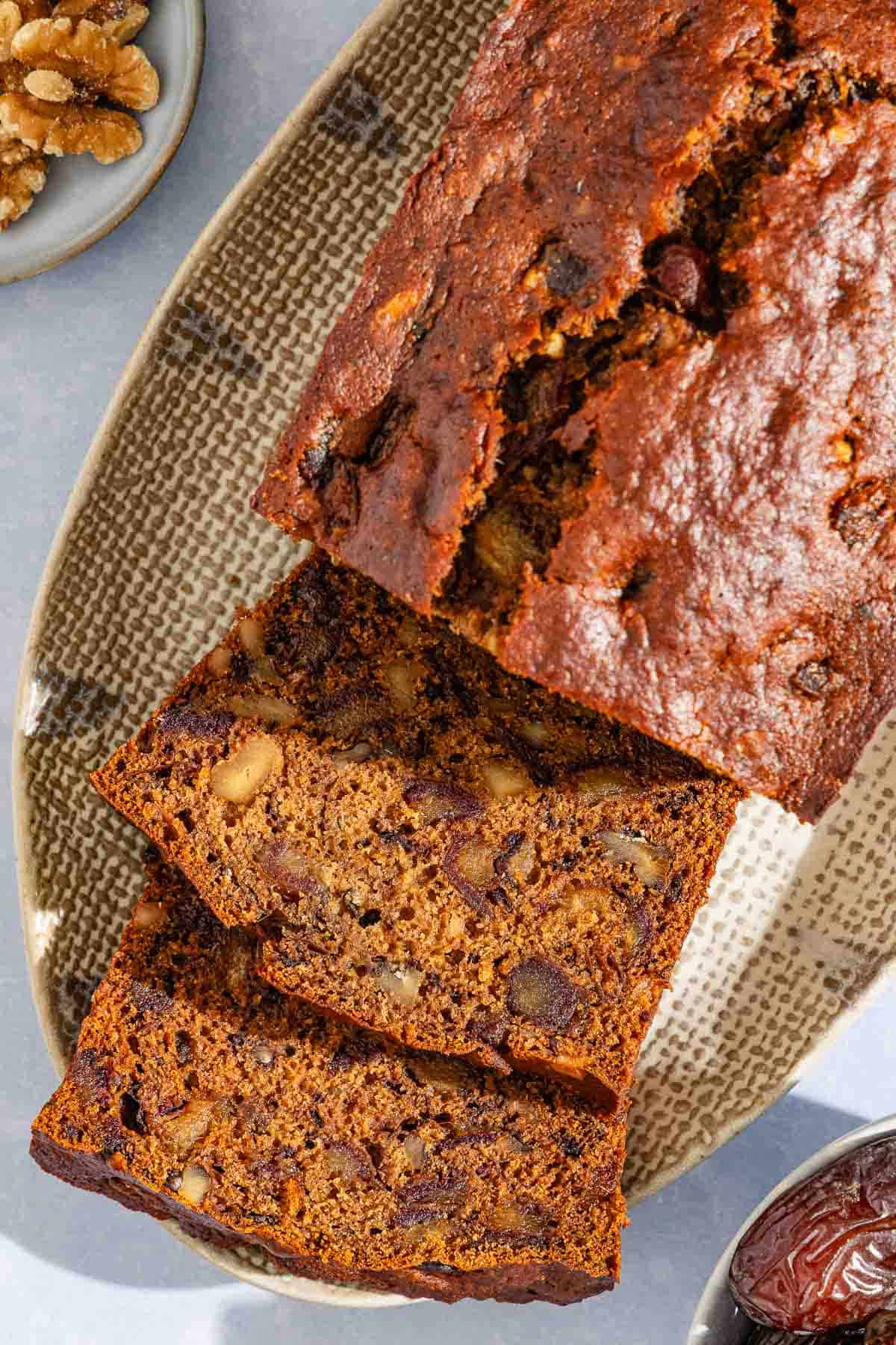 A partially sliced loaf of date nut bread behind 2 slices of the bread on a platter. Next to this are bowls of walnuts and dates.