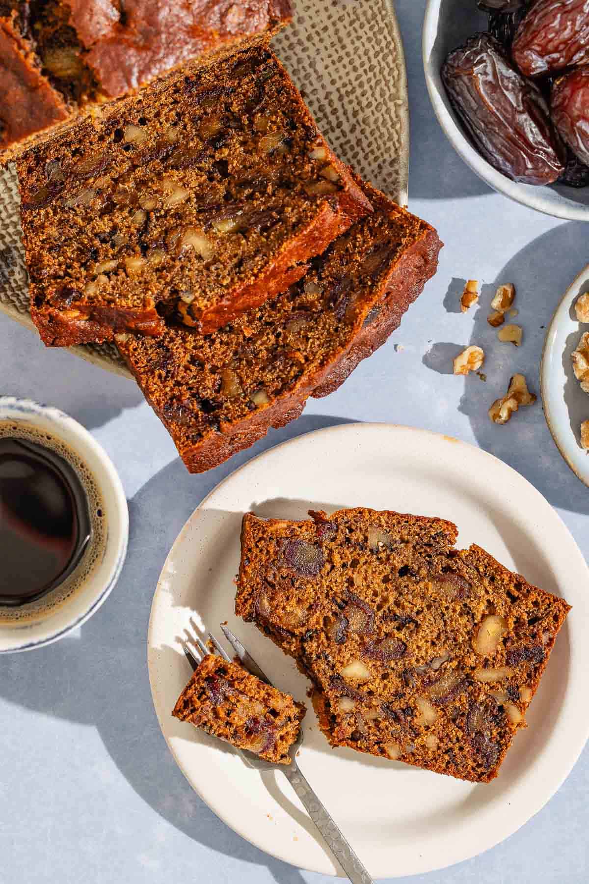 A slice of the date nut bread on a plate with a fork. Next to this is a cup of coffee, the rest of the bread on a platter, and bowls of dates and walnuts.