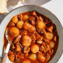 A serving of mushroom stew in a bowl with a spoon next to pieces of crusty bread and a glass of red wine.
