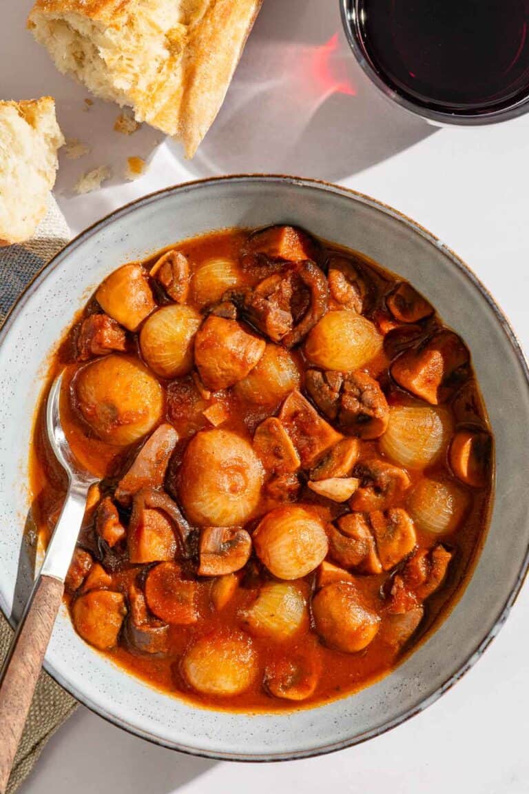 A serving of mushroom stew in a bowl with a spoon next to pieces of crusty bread and a glass of red wine.