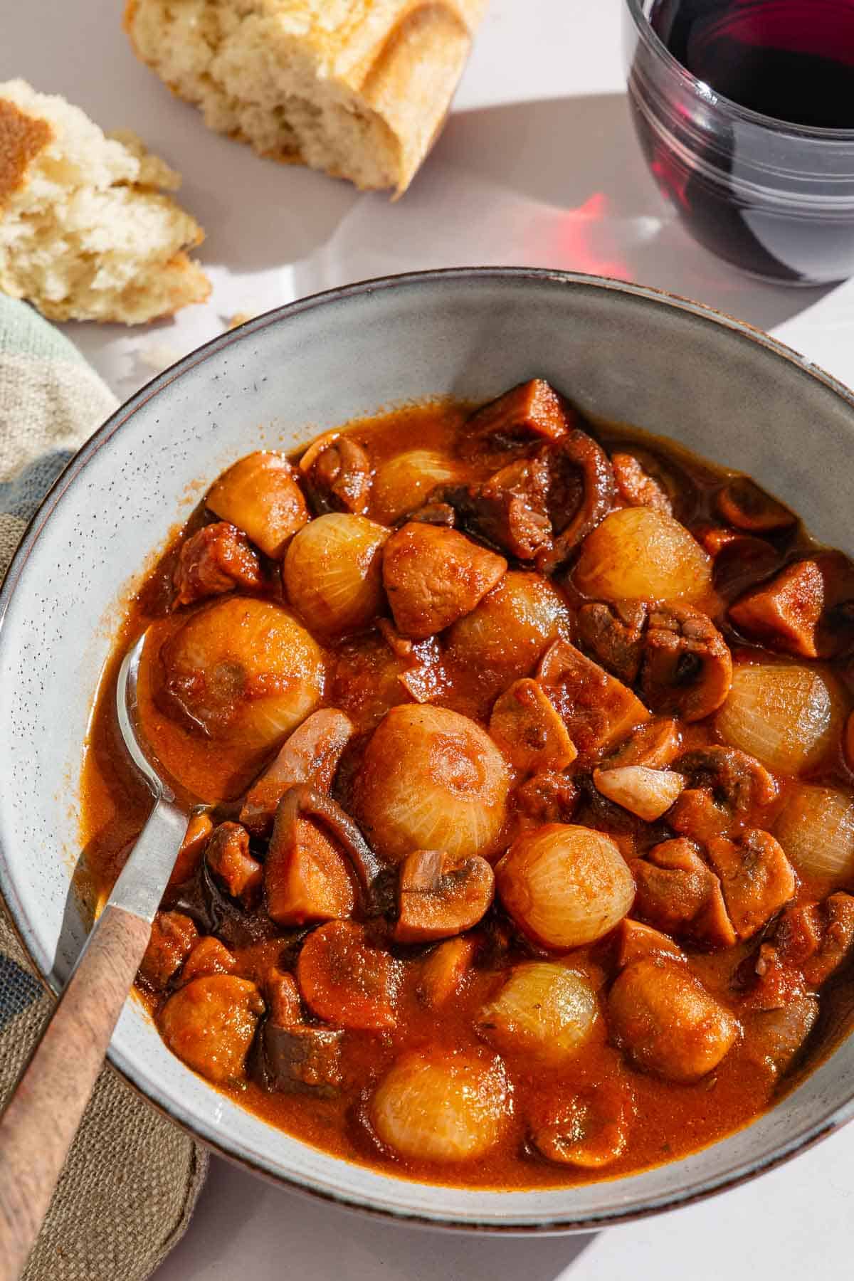 A serving of mushroom stew in a bowl with a spoon next to pieces of crusty bread and a glass of red wine.