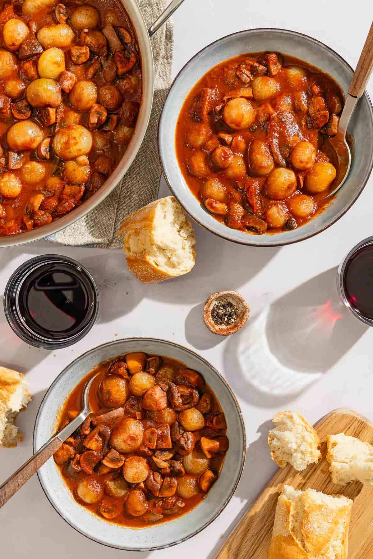 Two servings of the mushroom stew in a bowls with spoons. Next to these are pieces of crusty bread, 2 glasses of red wine, a bowl of peppercorns, and the rest of the stew in a tall sided skillet.