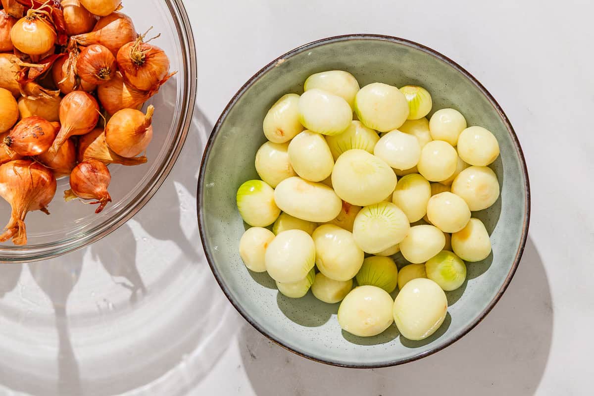 Peeled pearl onions in a bowl next to a bowl of unpeeled pearl onions.