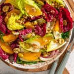 Orange salad in a serving bowl on a wooden tray with serving utensils. Next to this is a stack of 2 plates.