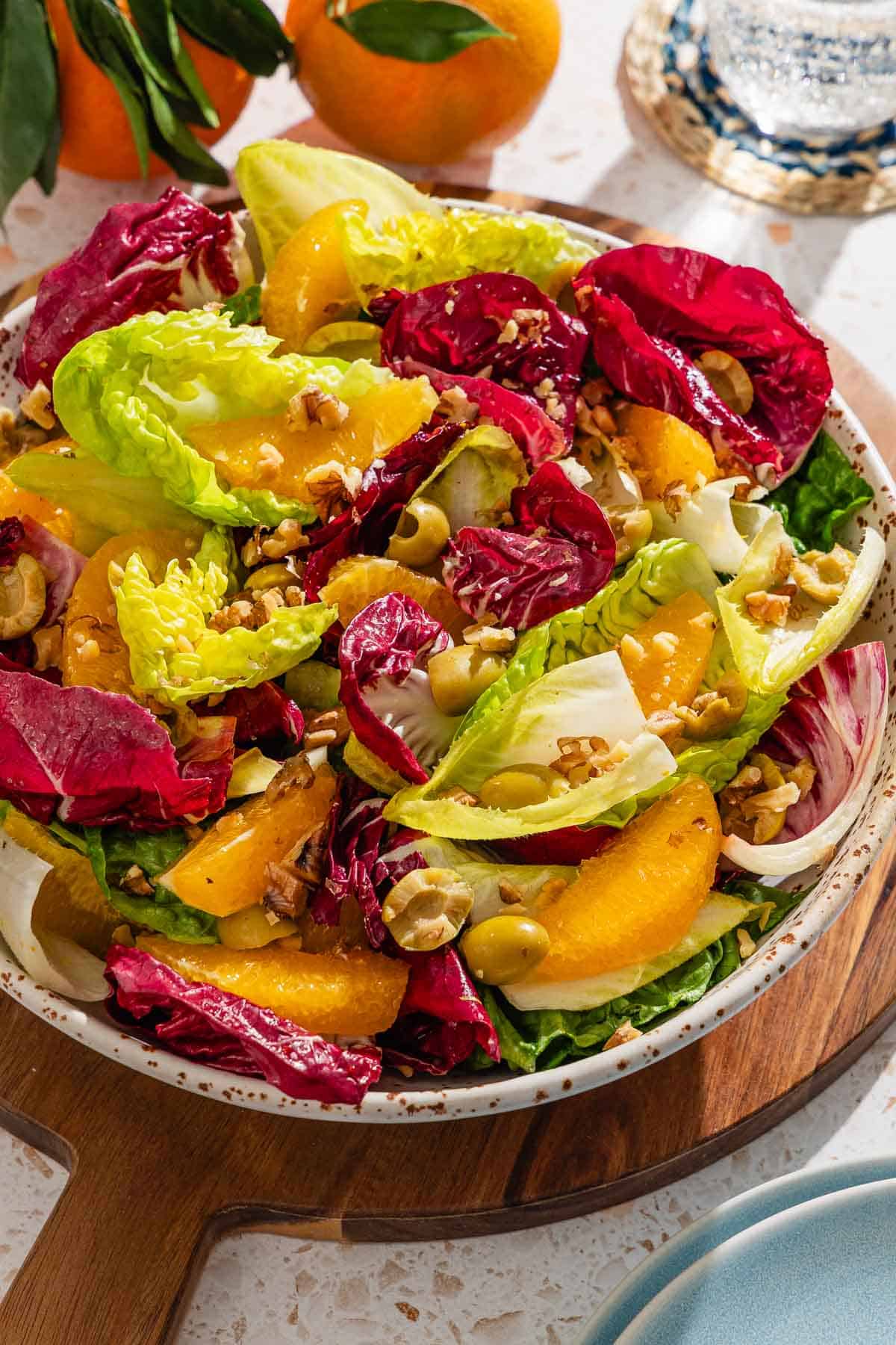 Orange salad in a serving bowl on a wooden tray. Next to this is a stack of 2 plates, a glass of sparkling water and 2 oranges.