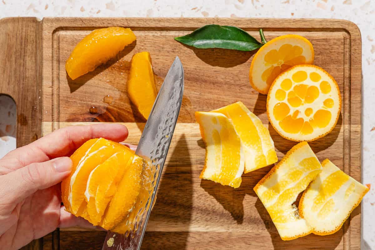 An orange being segmented with a knife on a cutting board. Also on the cutting board are the orange peels and 2 segments.