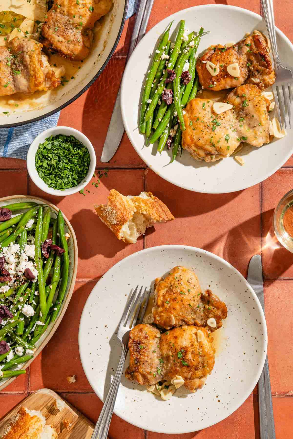 Two servings of the garlic chicken recipe on plates with forks. Next to these are knives, a green bean salad, a bowl of chopped parsley, a glass of white wine, pieces of crusty bread and the rest of the garlic chicken in a skillet.