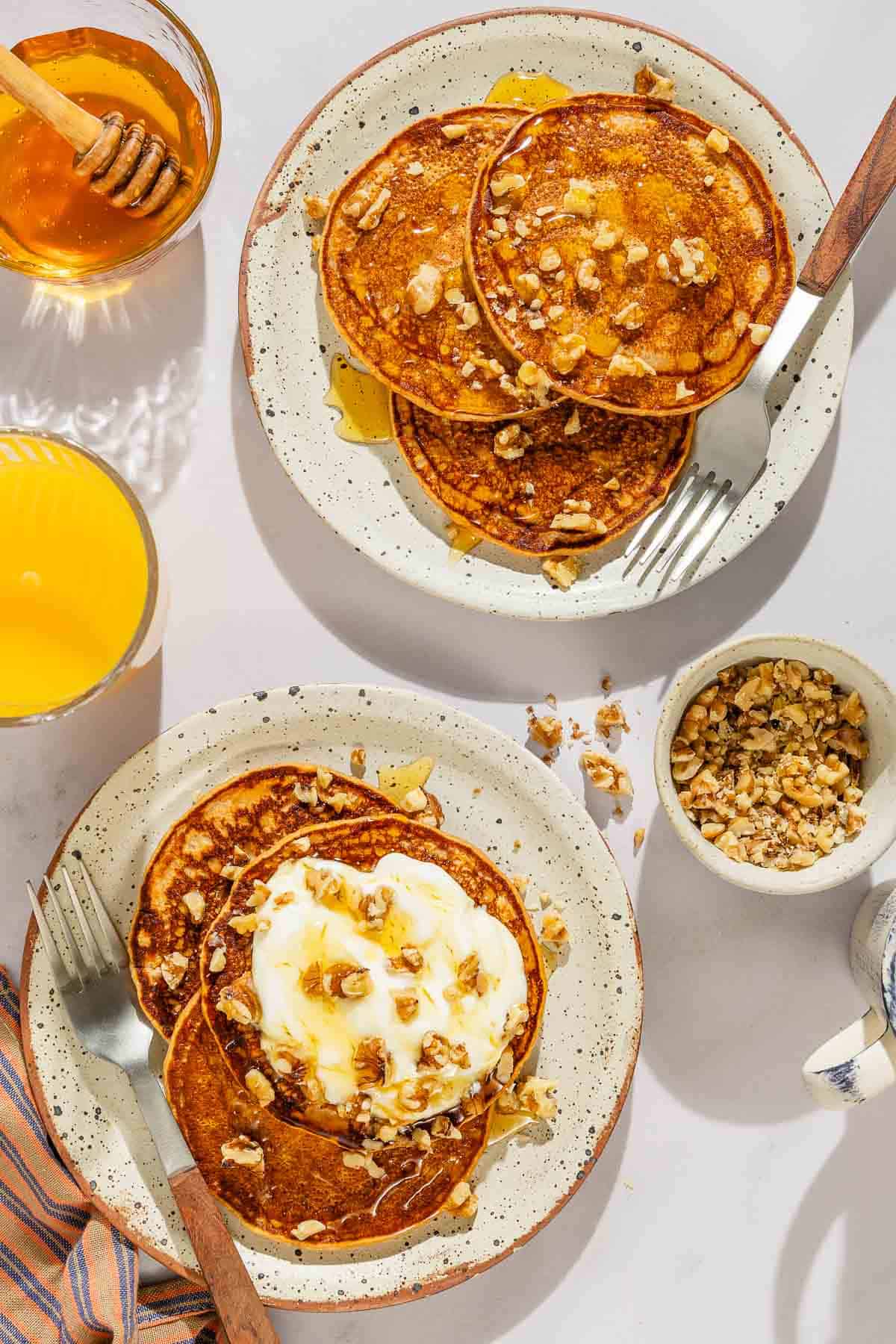 Two plates of the gluten free sweet potato pancakes with forks. Next to these are bowls of nuts and honey, and a glass of orange juice.