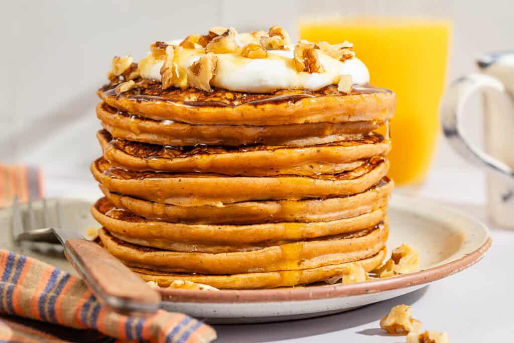 A stack of gluten free sweet potato pancakes topped with labneh, nuts and honey on a plate with a fork.