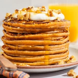A stack of gluten free sweet potato pancakes topped with labneh, nuts and honey on a plate with a fork.