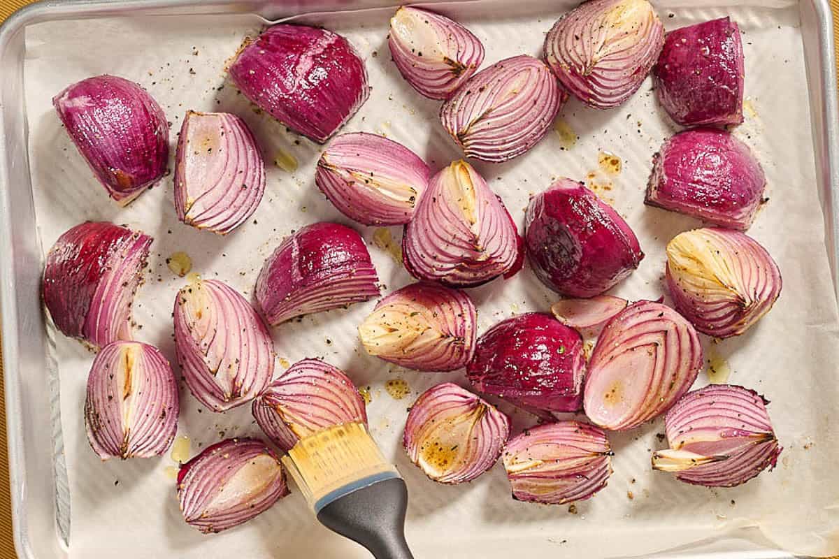 The partially cooked quartered red onions being basted with the balsamic vinaigrette on a parchment lined sheet pan.