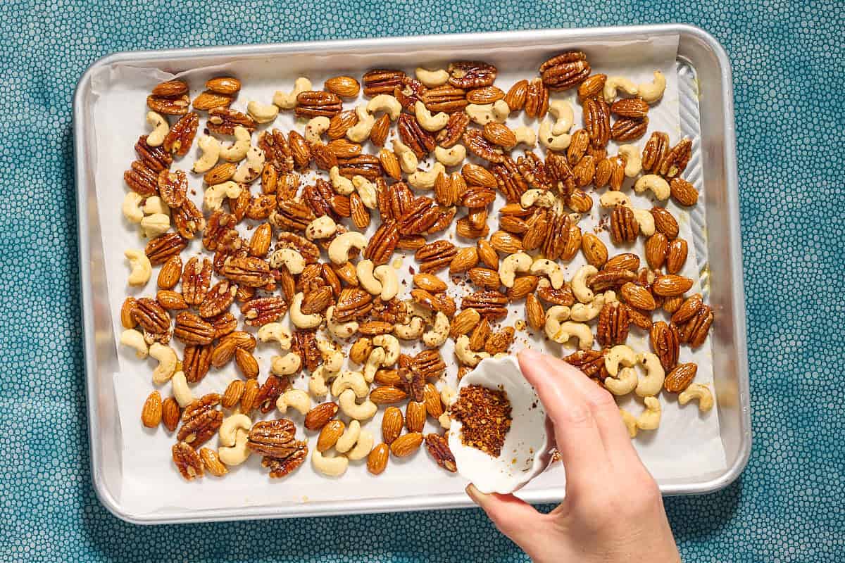 The seasonings being poured over the spiced nuts recipe on a parchment lined sheet pan.