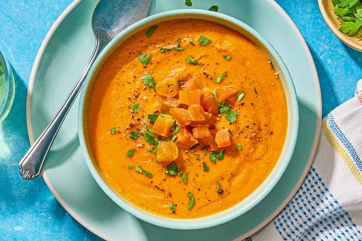 A bowl of red lentil soup topped with chopped dried apricots and parsley on a plate with a spoon.