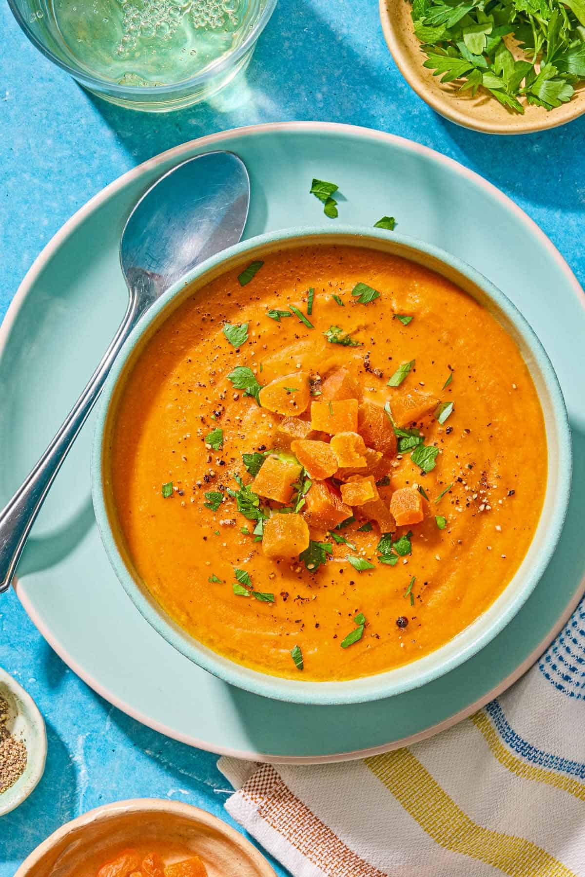 A bowl of red lentil soup topped with chopped dried apricots and parsley on a plate with a spoon and a piece of crusty bread. Next to this is a glass of white wine and bowls of chopped parsely and dried apricots.