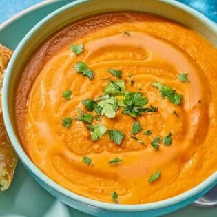 A bowl of red lentil soup topped with parsley on a plate with a spoon and a piece of crusty bread.