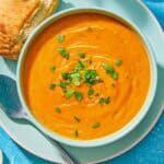 A bowl of red lentil soup topped with chopped dried apricots and parsley on a plate with a spoon and a piece of crusty bread.