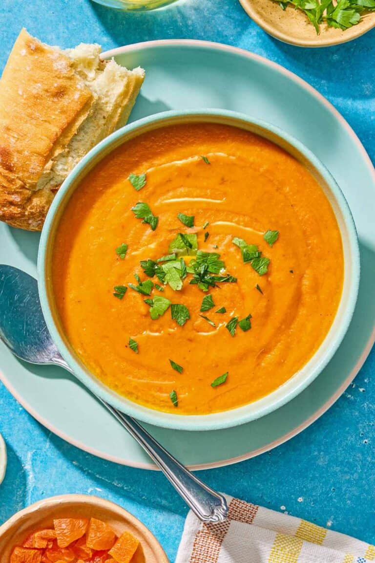 A bowl of red lentil soup topped with chopped dried apricots and parsley on a plate with a spoon and a piece of crusty bread.