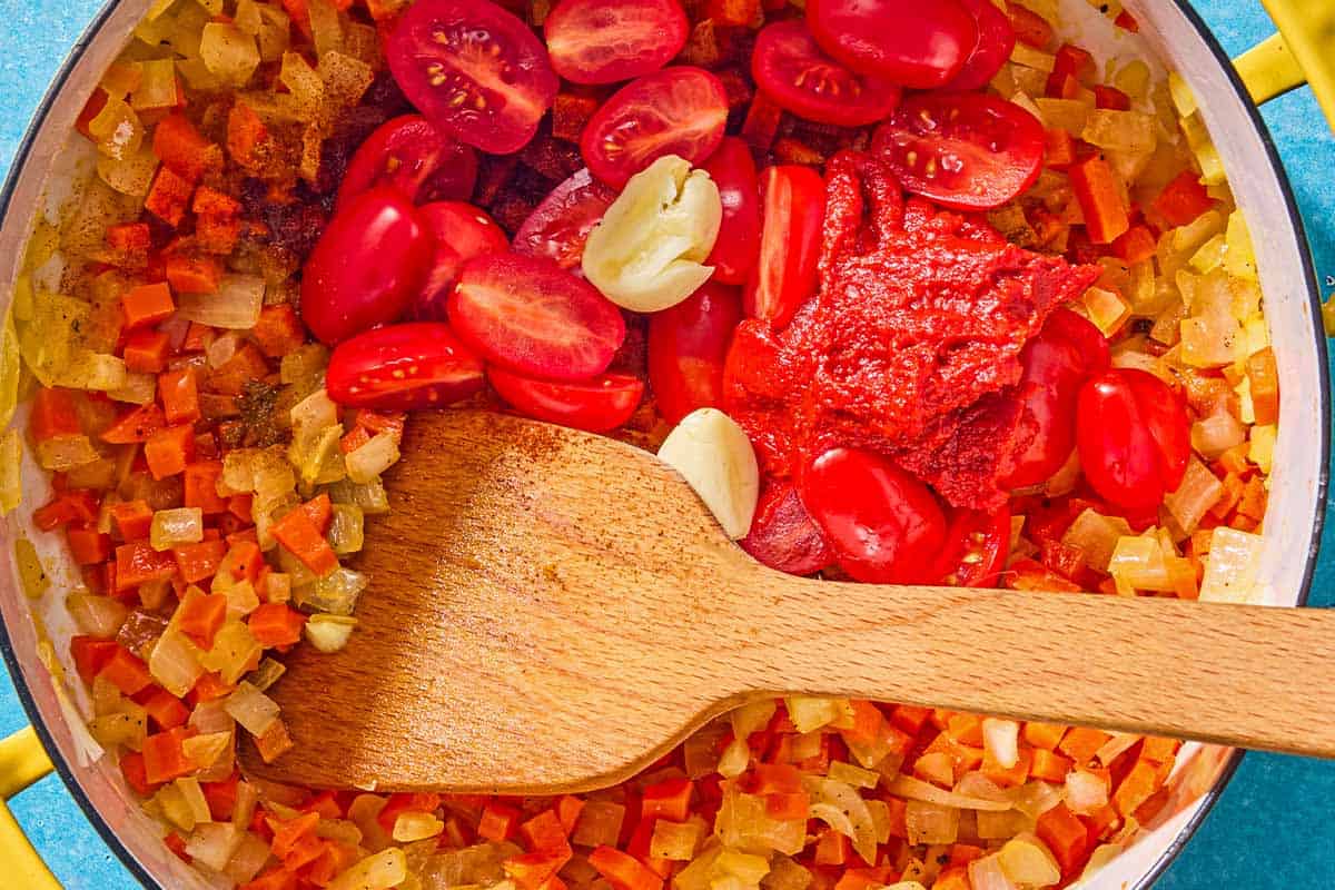 The cherry tomatoes, tomato paste and garlic being stirred into the sauteed vegetables with a wooden spoon in a pot.