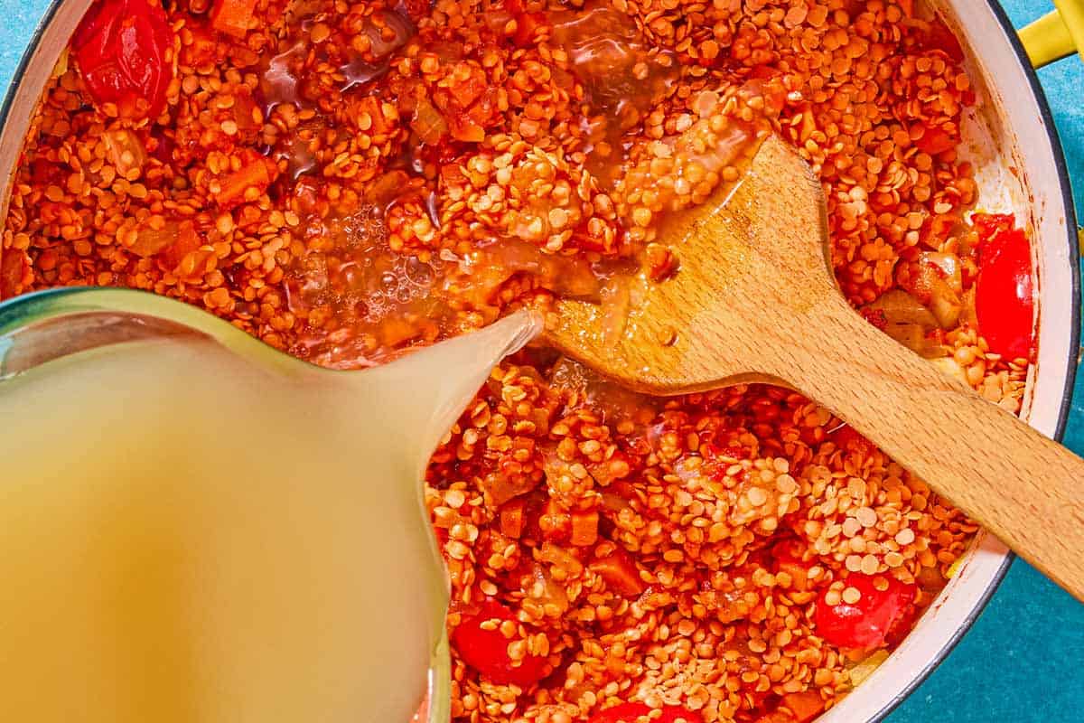 The chicken stock being added to the rest of the ingredients for the red lentil soup in a large pot with a wooden spoon.