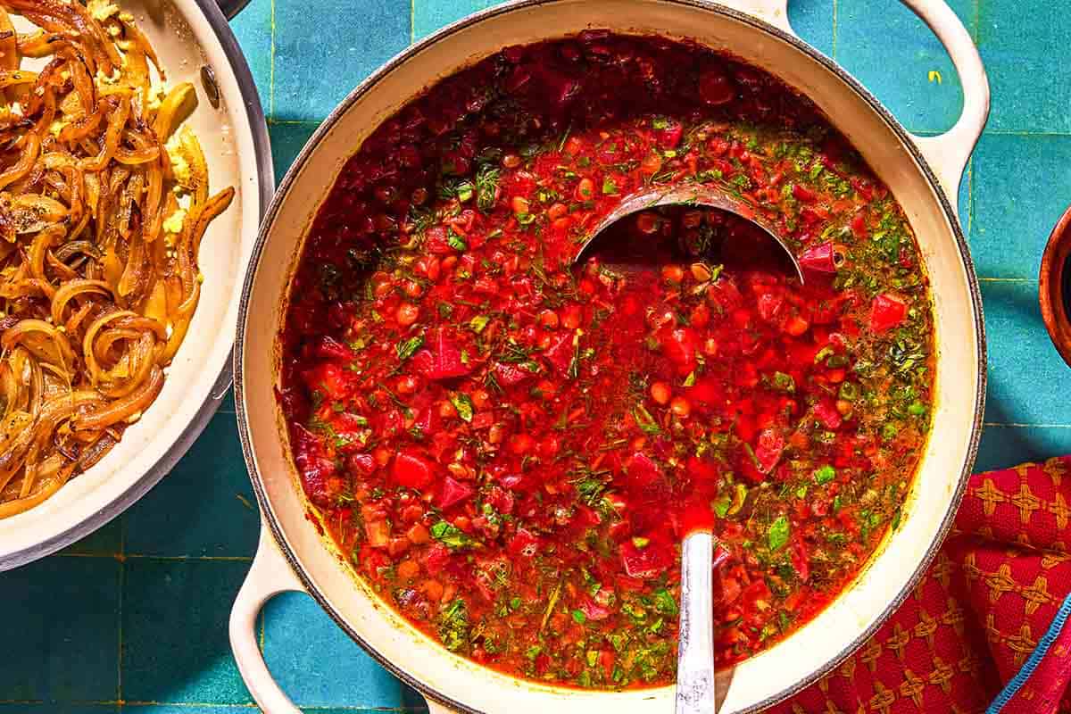 Beet soup in a pot with a ladle next to a skillet with fried onions.