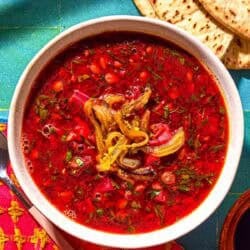 A bowl of beet soup topped with fried onions. Next to this is a spoon, a bowl of pomegranate molasses and pita bread.
