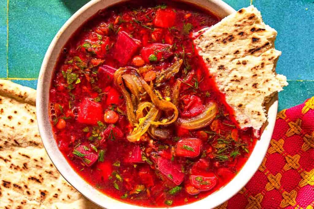 A bowl of beet soup topped with fried onions and a piece of pita bread next to more pita bread.