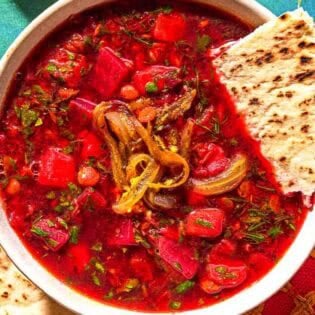 A bowl of beet soup topped with fried onions and a piece of pita bread next to more pita bread.