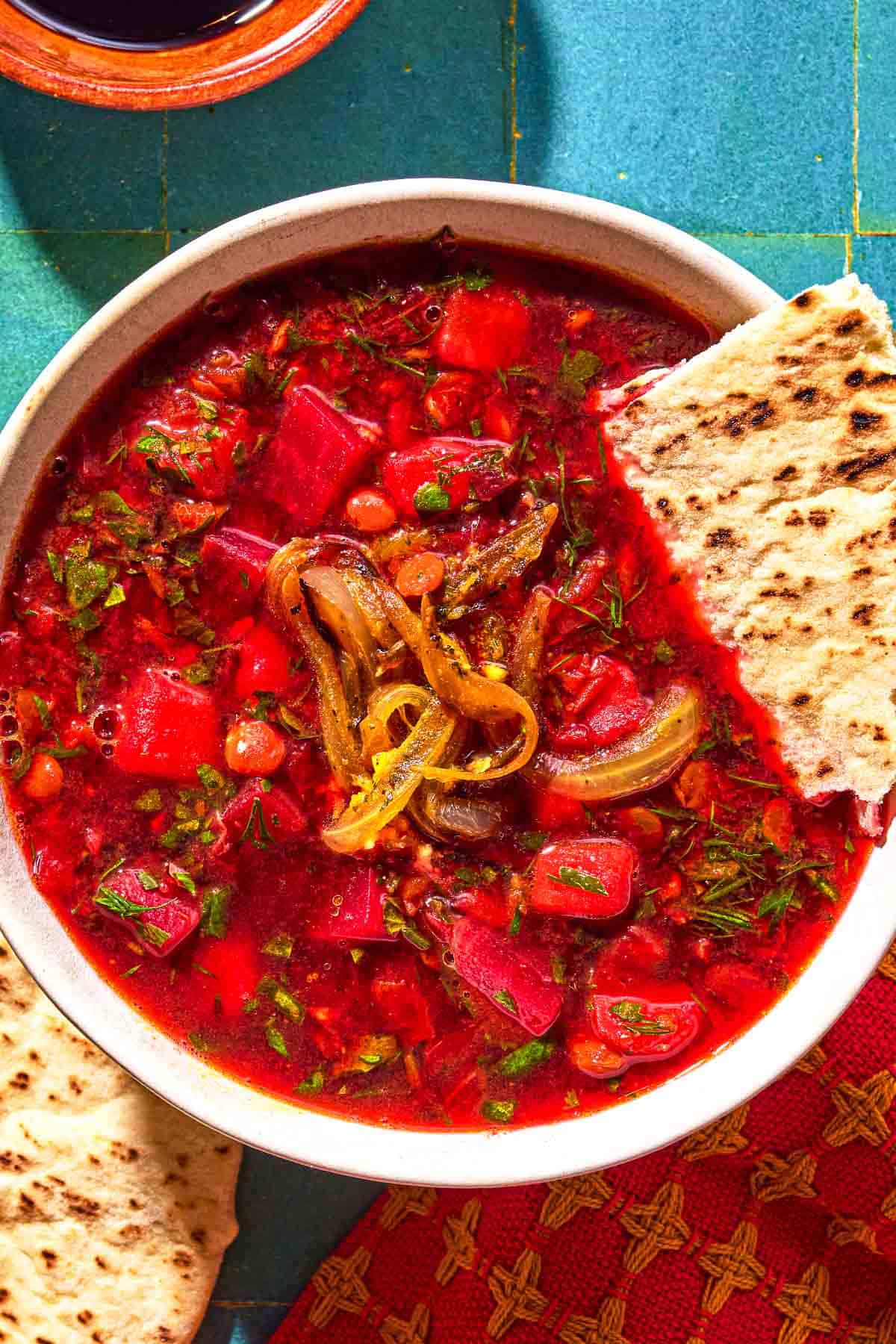 A bowl of Persian beet soup (Ash-e Shooli) topped with fried onions and a piece of pita bread. Next to this is a bowl of pomegranate molasses and another piece of pita bread.