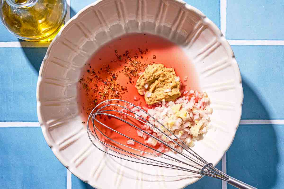 The ingredients for the red wine vinaigrette just before being mixed together in a bowl with a whisk next to a bottle of olive oil.
