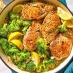 The chicken and broccoli in a skillet with lemon wedges next to a kitchen towel and a bowl of oregano.
