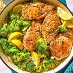The chicken and broccoli in a skillet with lemon wedges next to a kitchen towel and a bowl of oregano.