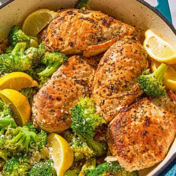 The chicken and broccoli in a skillet with lemon wedges next to a kitchen towel and a bowl of Parmesan cheese.