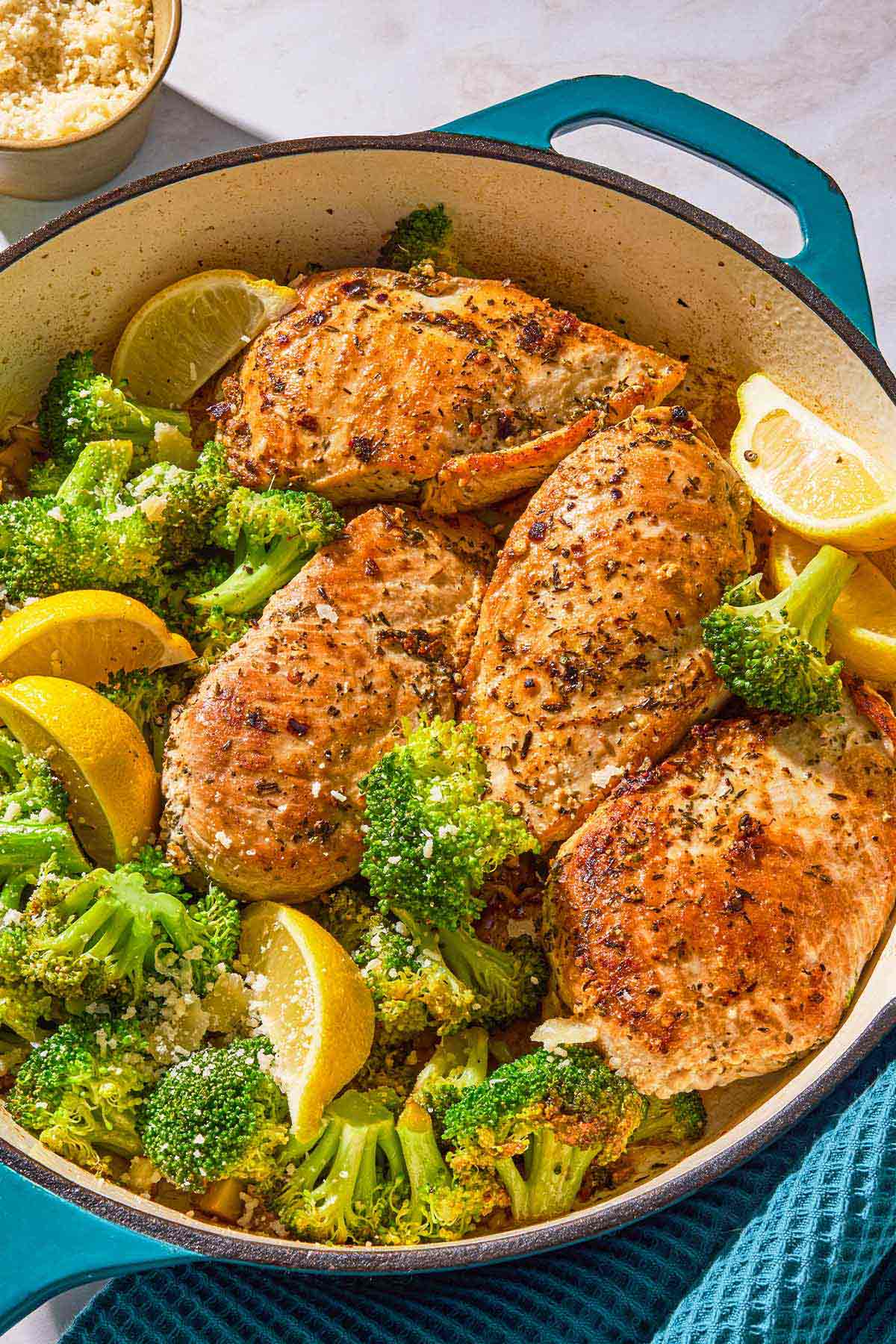 The chicken and broccoli in a skillet with lemon wedges next to a kitchen towel and a bowl of Parmesan cheese.
