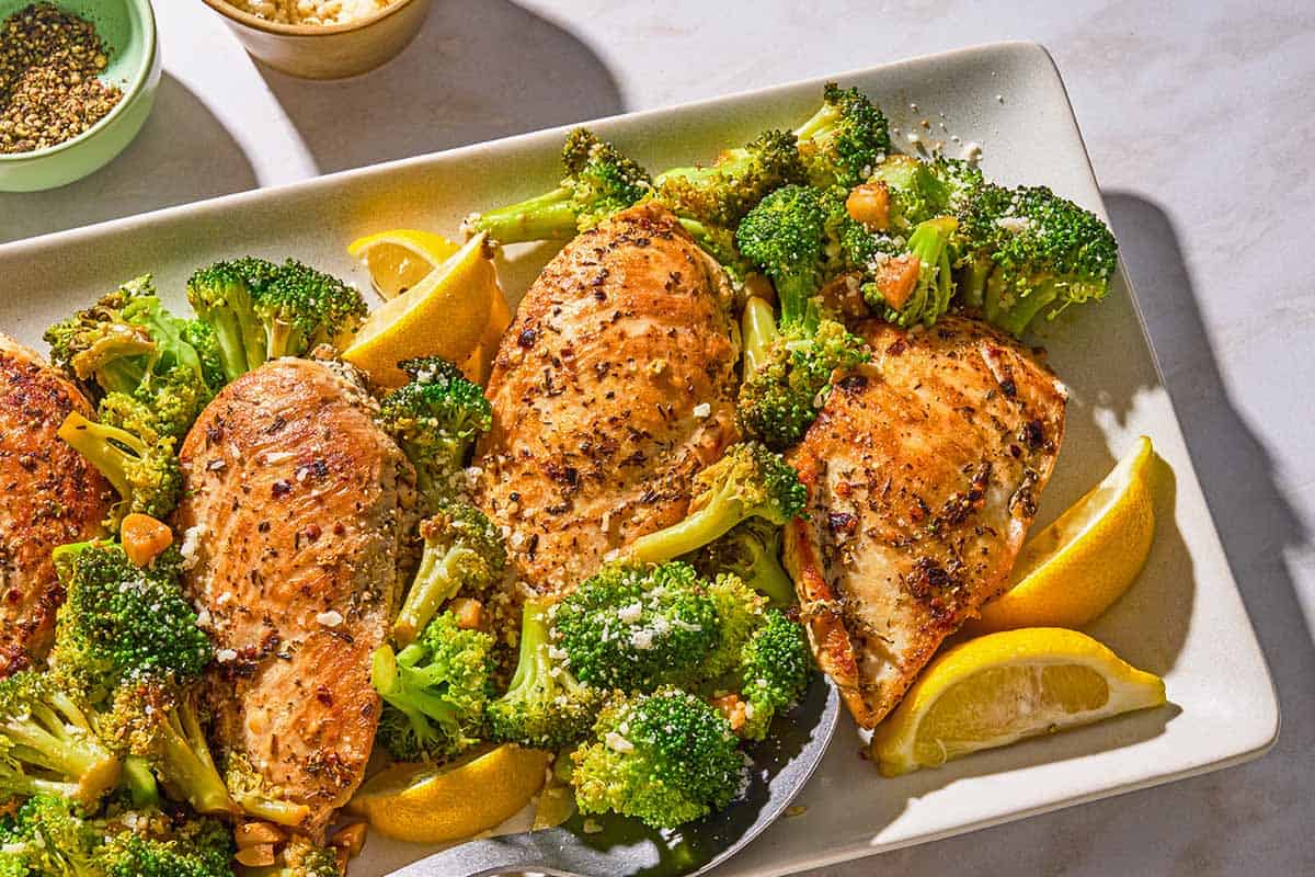 Chicken and broccoli on a platter with lemon wedges next to bowls of oregano and grated Parmesan cheese.