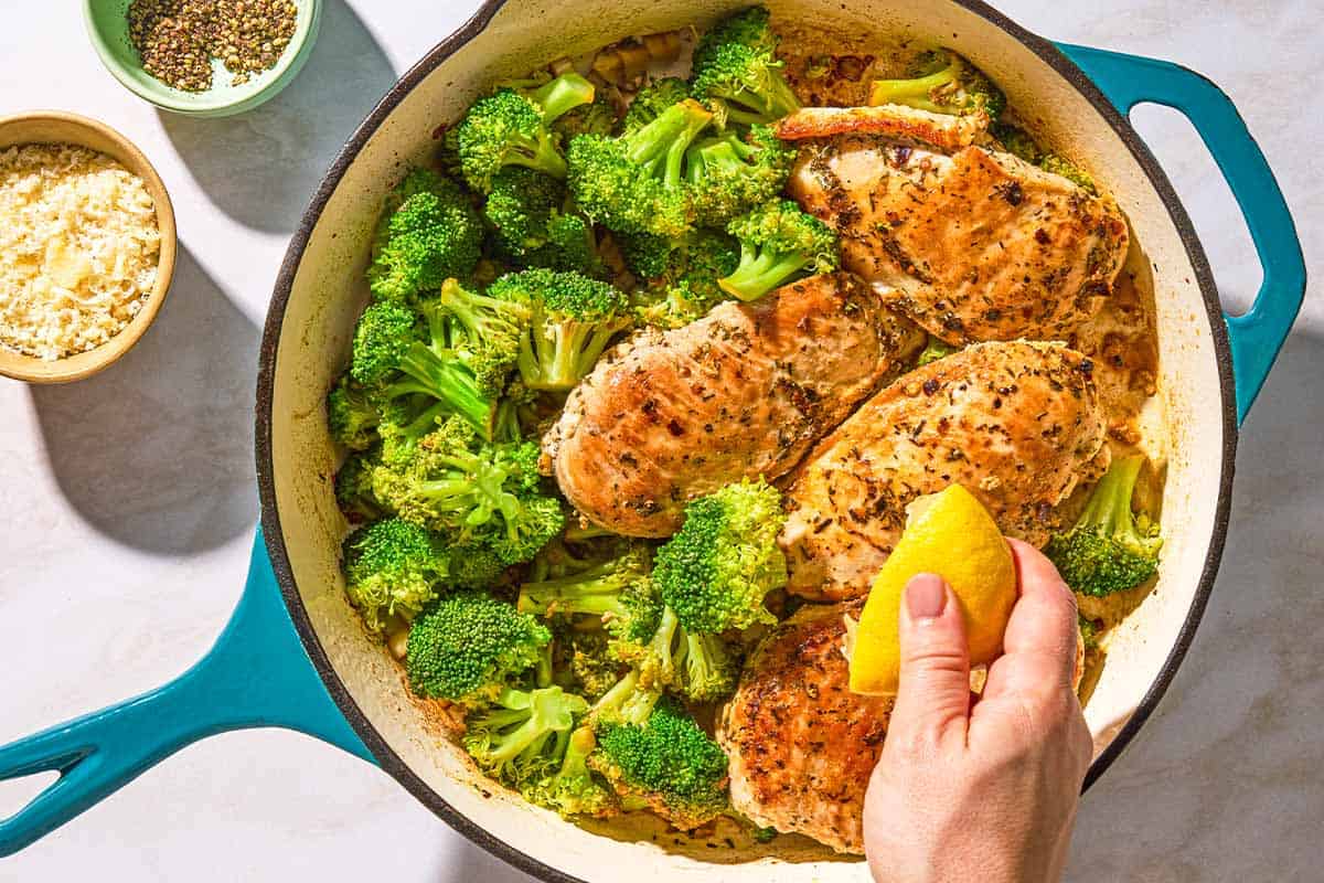 A lemon half being squeezed over the chicken and broccoli in a skillet. Next to this are bowls of oregano and grated parmesan cheese.