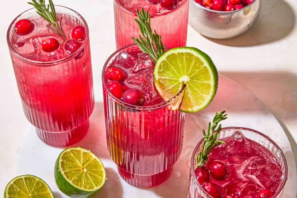 Several glasses of cranberry mocktail garnished with rosemary, cranberries and lime. Next to these are more limes, and a bowl of cranberries.