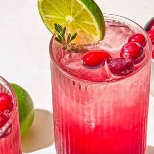 Two glasses of the cranberry mocktail garnished with rosemary, cranberries and lime next to a lime and a bowl of cranberries.