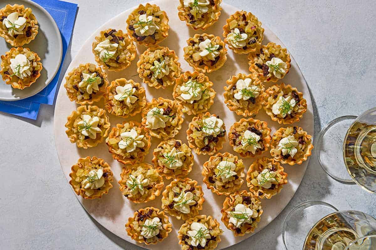 Several phyllo cups spread evenly on a platter next to a plate with more phyllo cups and two glasses of white wine.