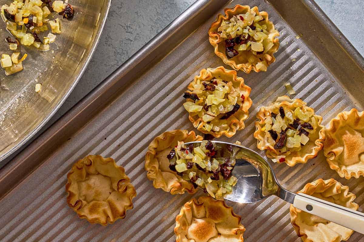 Filling being added to phyllo cups on a sheet pan with a spoon.
