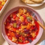 A serving of oven roasted vegetables in a bowl next to a kitchen towel, a plate with pita and the rest of the vegetables in a baking dish.