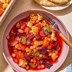 A serving of oven roasted vegetables in a bowl next to a kitchen towel, a plate with pita and the rest of the vegetables in a baking dish.