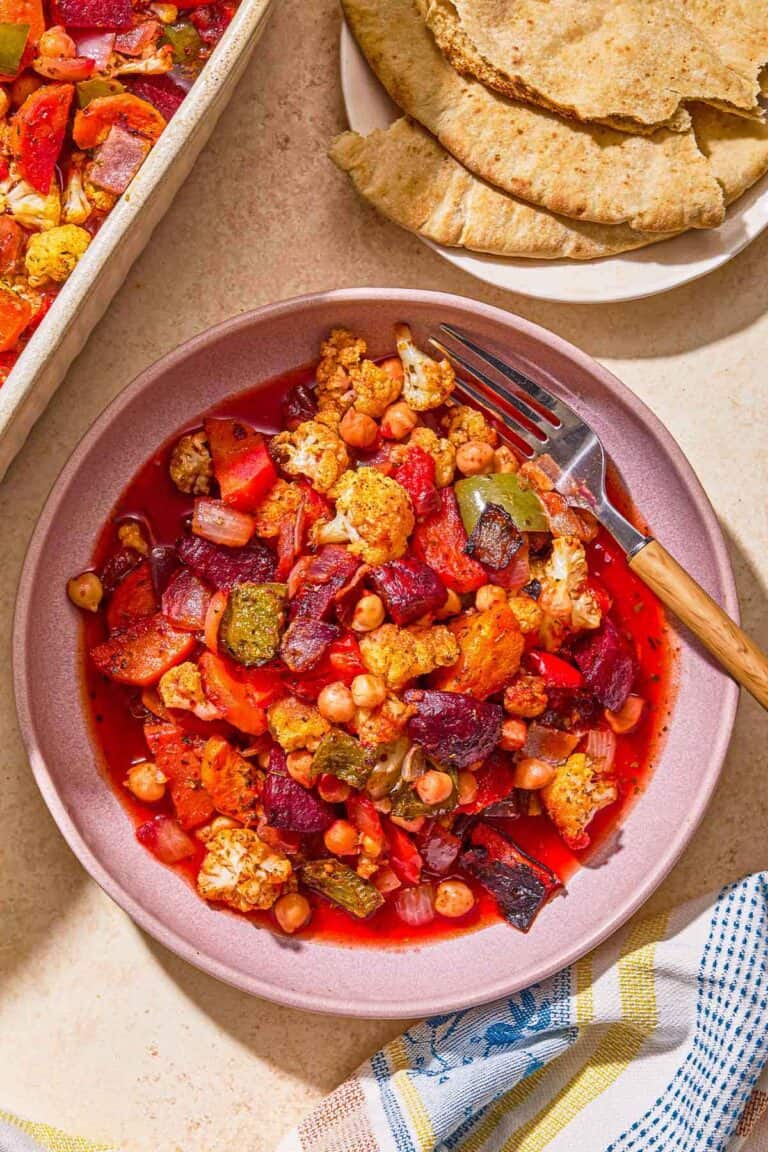 A serving of oven roasted vegetables in a bowl next to a kitchen towel, a plate with pita and the rest of the vegetables in a baking dish.