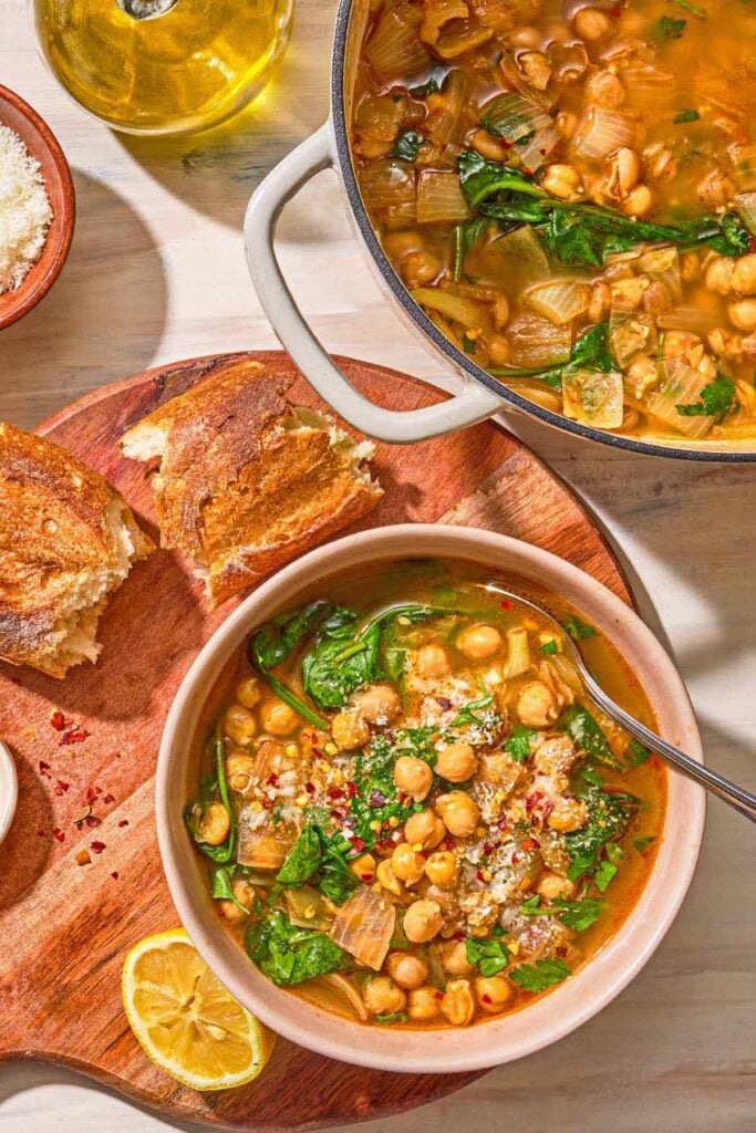 A serving of spinach soup in a bowl with a spoon. Next to this is a lemon half, pieces of crusty bread, a bowl of pecorino Romano cheese, a bottle of olive oil, and the rest of the soup in a pot.